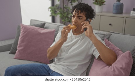 A young hispanic man multitasking, eating while talking on the phone in a modern indoor setting. - Powered by Shutterstock