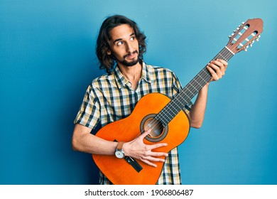 Young hispanic man playing classical guitar smiling looking to the side and staring away thinking. 