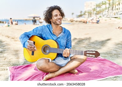 Young hispanic man playing classical guitar sitting on sand at the beach.