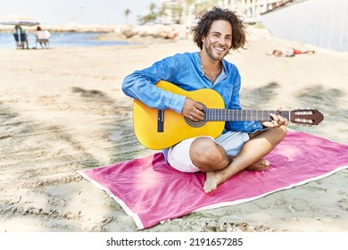 Young hispanic man playing classical guitar sitting on sand at the beach.