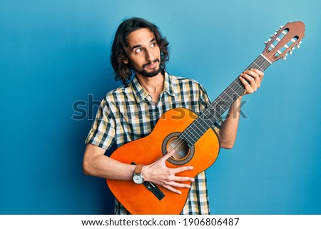 Young hispanic man playing classical guitar smiling looking to the side and staring away thinking. 