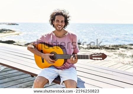 Young hispanic man playing classical guitar sitting on  bench at the beach.