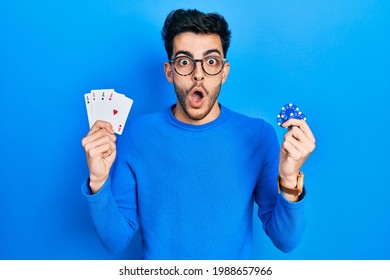 Young hispanic man playing poker holding casino chips and cards afraid and shocked with surprise and amazed expression, fear and excited face.  - Powered by Shutterstock