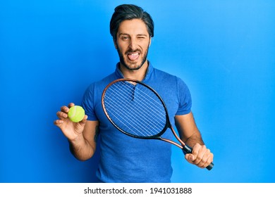 Young Hispanic Man Playing Tennis Holding Racket And Ball Sticking Tongue Out Happy With Funny Expression. 