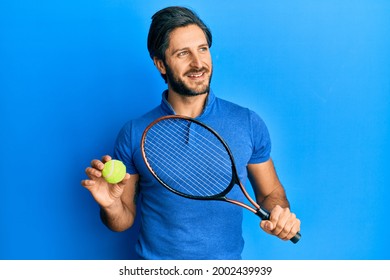 Young Hispanic Man Playing Tennis Holding Racket And Ball Smiling Looking To The Side And Staring Away Thinking. 