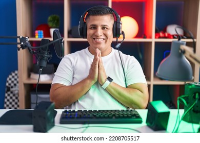 Young Hispanic Man Playing Video Games Praying With Hands Together Asking For Forgiveness Smiling Confident. 