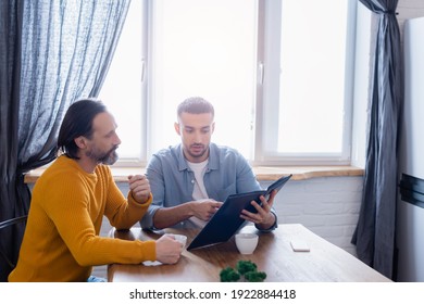 Young Hispanic Man Pointing At Photo Album Near Bearded Father In Kitchen