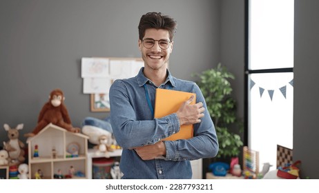 Young hispanic man preschool teacher smiling confident holding book at kindergarten - Powered by Shutterstock