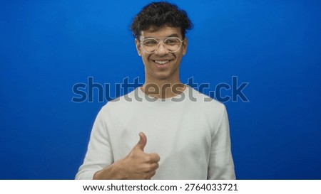 Young hispanic man presenting open palm and smiling with glasses in blue studio; friendly greeting welcome.