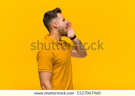 young hispanic man profile view, looking happy and excited, shouting and calling to copy space on the side against orange wall