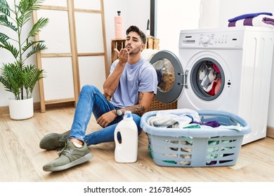 Young Hispanic Man Putting Dirty Laundry Into Washing Machine Looking At The Camera Blowing A Kiss With Hand On Air Being Lovely And Sexy. Love Expression. 