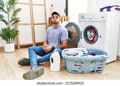 Young Hispanic Man Putting Dirty Laundry Into Washing Machine Puffing Cheeks With Funny Face. Mouth Inflated With Air, Crazy Expression. 