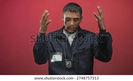 Young hispanic man raises hands showing police badge and idcard against red backdrop in studio; contemplation.