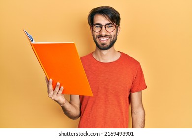 Young Hispanic Man Reading Book Looking Positive And Happy Standing And Smiling With A Confident Smile Showing Teeth 