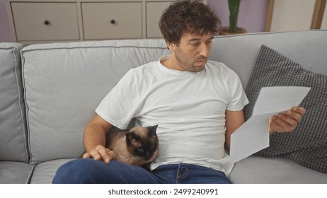 Young hispanic man reading a document with a siamese cat sitting beside him on a couch indoors.