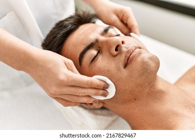 Young hispanic man relaxed having facial treatment cleaning face with cotton disk at beauty center