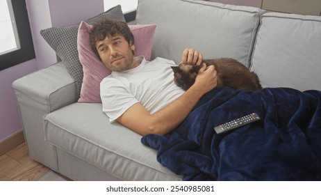 A young hispanic man relaxes with his pet cat on a sofa in the living room, watching tv indoors with a remote in his hand. - Powered by Shutterstock