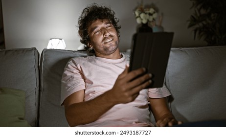 A young hispanic man relaxes at home using a tablet on a couch in a dimly lit living room. - Powered by Shutterstock