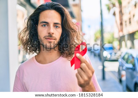Young hispanic man with serious expression holding hiv awareness red ribbon at city.