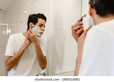 Young Hispanic Man Shaving His Face Standing At The Mirror Indoors