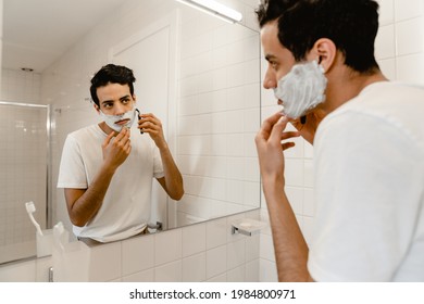 Young Hispanic Man Shaving His Face Standing At The Mirror Indoors