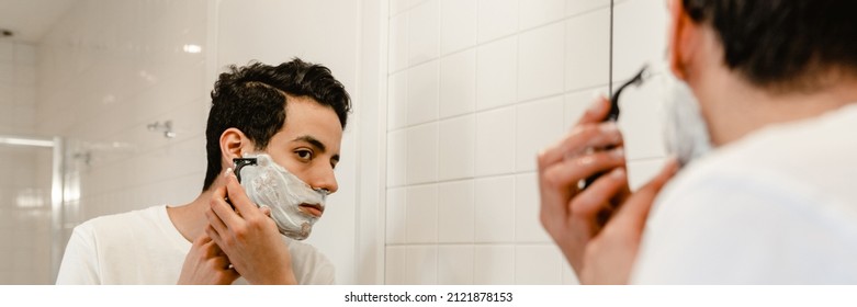 Young Hispanic Man Shaving His Face Standing At The Mirror Indoors