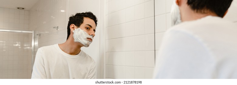 Young Hispanic Man Shaving His Face Standing At The Mirror Indoors