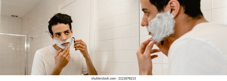 Young Hispanic Man Shaving His Face Standing At The Mirror Indoors