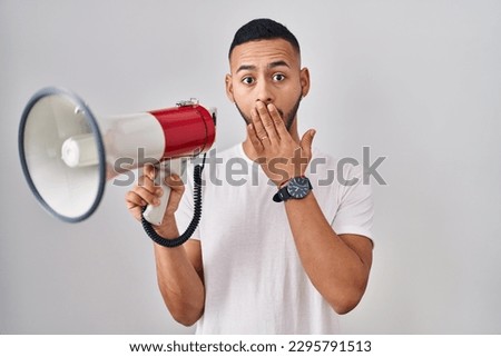 Young hispanic man shouting through megaphone covering mouth with hand, shocked and afraid for mistake. surprised expression 