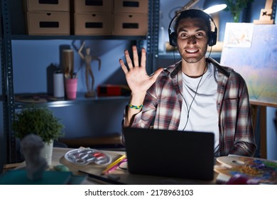 Young Hispanic Man Sitting At Art Studio With Laptop Late At Night Showing And Pointing Up With Fingers Number Five While Smiling Confident And Happy. 