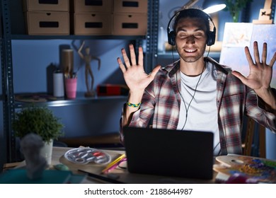 Young Hispanic Man Sitting At Art Studio With Laptop Late At Night Showing And Pointing Up With Fingers Number Ten While Smiling Confident And Happy. 