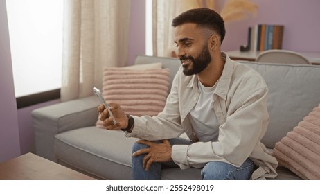 Young hispanic man sitting in a cozy living room using a smartphone, wearing casual clothes and smiling while relaxed at home. - Powered by Shutterstock