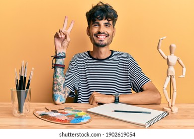 Young hispanic man sitting on the table at art studio smiling with happy face winking at the camera doing victory sign. number two. 