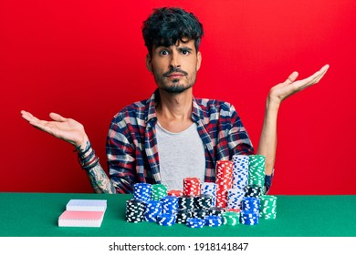 Young Hispanic Man Sitting On The Table With Poker Chips And Cards Clueless And Confused With Open Arms, No Idea And Doubtful Face. 