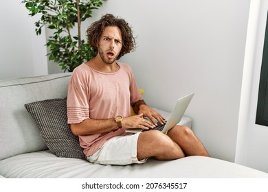 Young hispanic man sitting on the sofa at home using laptop in shock face, looking skeptical and sarcastic, surprised with open mouth  - Shutterstock ID 2076345517