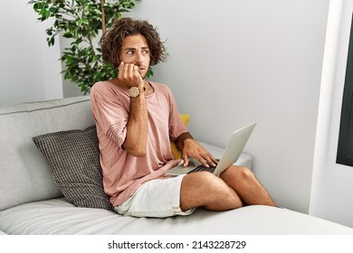 Young Hispanic Man Sitting On The Sofa At Home Using Laptop Looking Stressed And Nervous With Hands On Mouth Biting Nails. Anxiety Problem. 