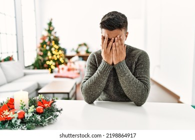 Young Hispanic Man Sitting On The Table By Christmas Tree With Sad Expression Covering Face With Hands While Crying. Depression Concept. 