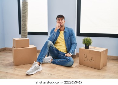 Young Hispanic Man Sitting On The Floor At New Home With Hand On Chin Thinking About Question, Pensive Expression. Smiling With Thoughtful Face. Doubt Concept. 