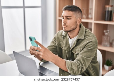 Young hispanic man sitting on table using smartphone at home
