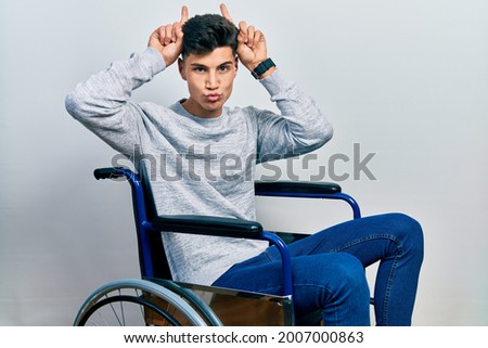 Young hispanic man sitting on wheelchair doing funny gesture with finger over head as bull horns 
