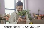 A young hispanic man smiles while smelling a potted plant inside a colorful flower shop.