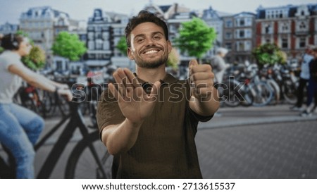 Young hispanic man smiling with bare hands forward miming a steering wheel on a busy street in amsterdam beside parked bicycles; joy travel exploration.