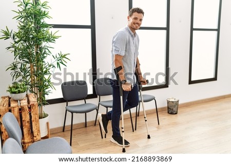 Young hispanic man smiling confident walking using crutches at clinic waiting room