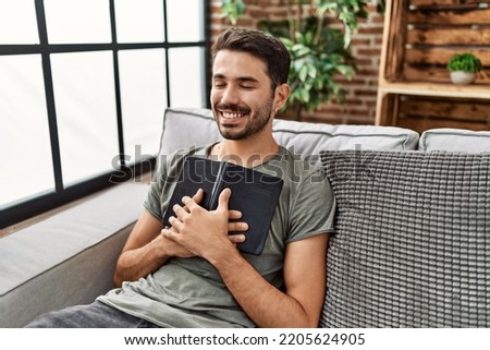 Young hispanic man smiling confident hugging bible at home