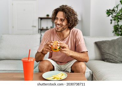 Young Hispanic Man Smiling Happy Eating Classic Burger And Drinking Soda At Home.