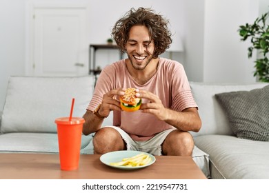 Young Hispanic Man Smiling Happy Eating Classic Burger And Drinking Soda At Home.