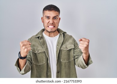 Young Hispanic Man Standing Over Isolated Background Angry And Mad Raising Fists Frustrated And Furious While Shouting With Anger. Rage And Aggressive Concept. 