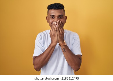 Young Hispanic Man Standing Over Yellow Background Laughing And Embarrassed Giggle Covering Mouth With Hands, Gossip And Scandal Concept 