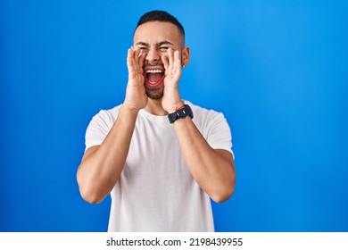 Young hispanic man standing over blue background shouting angry out loud with hands over mouth 