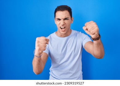 Young hispanic man standing over blue background angry and mad raising fists frustrated and furious while shouting with anger. rage and aggressive concept.  - Shutterstock ID 2309319527